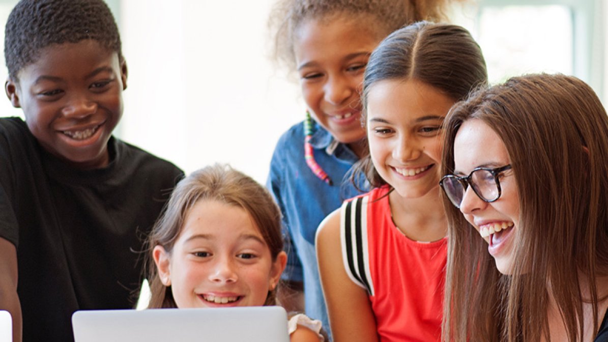 Young female teacher with pupils gathered around a laptop